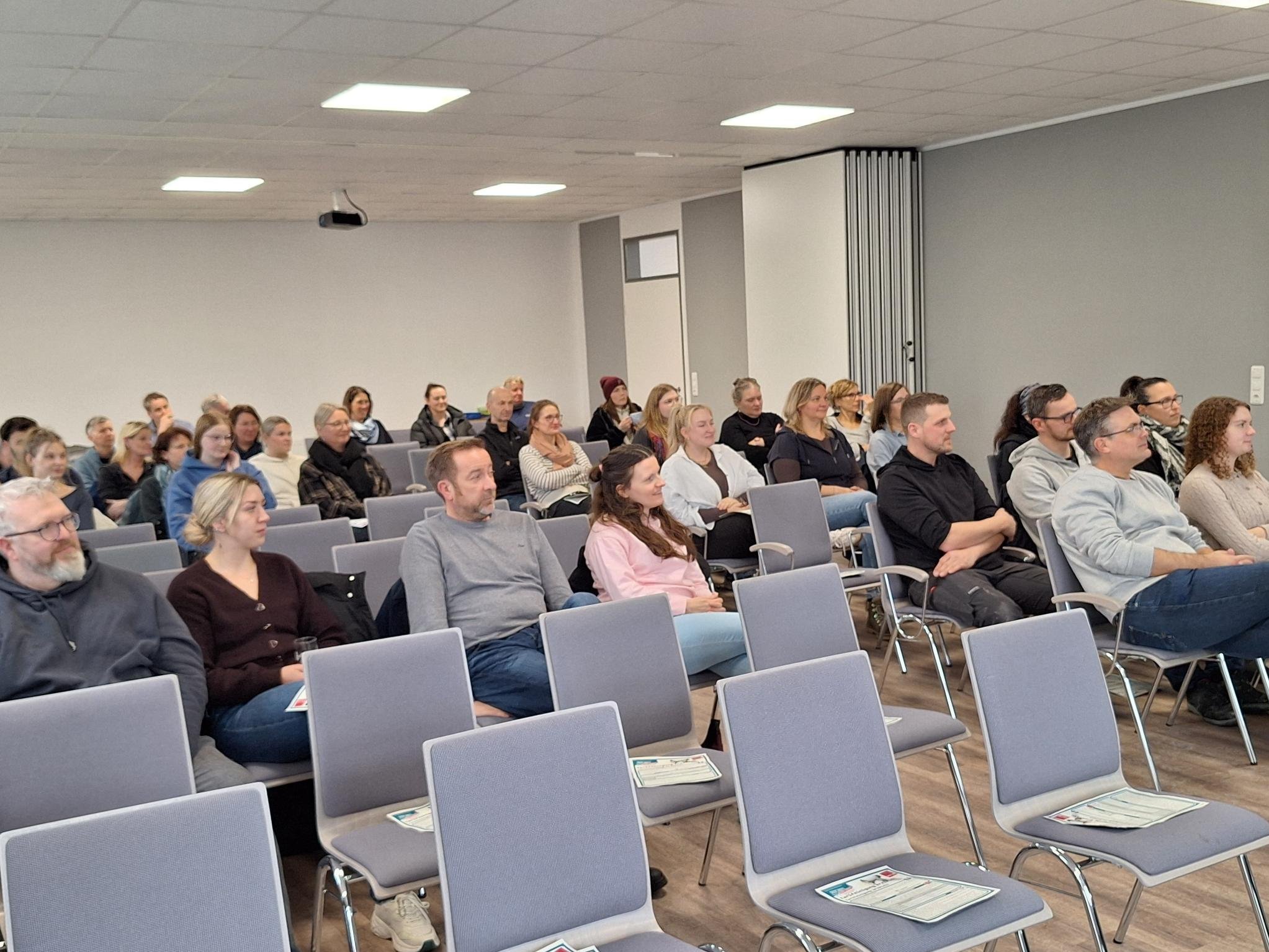Mehrere Mitarbeitende der Jugendhilfe Collstede sitzen in einem Veranstaltungsraum auf Stuhlreihen und verfolgen aufmerksam eine Präsentation während der Mitarbeitendenversammlung.