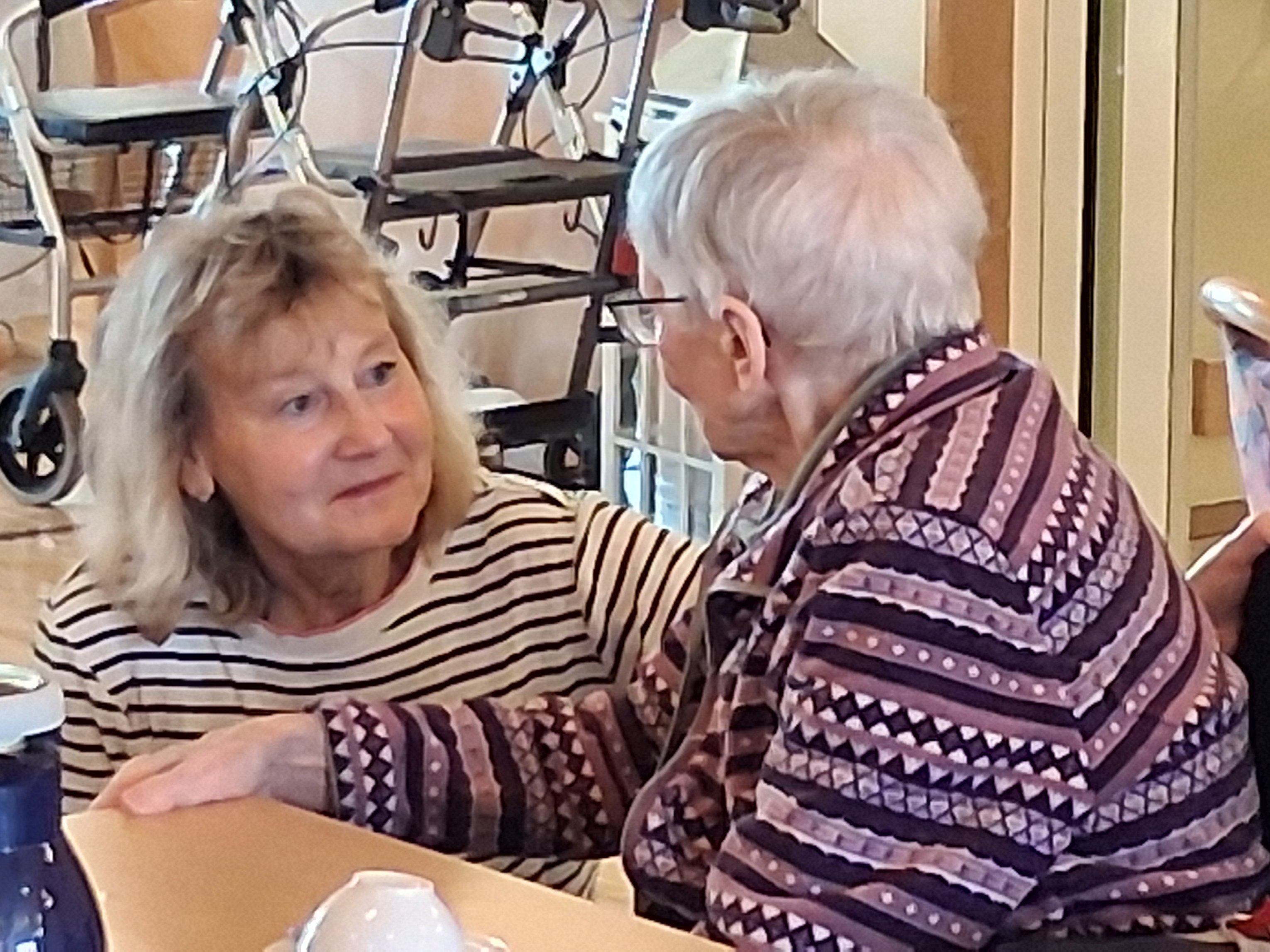 Eine Frau in einem gestreiften Oberteil unterhält sich zugewandt mit einer älteren Frau im gemusterten Pullover an einem Kaffeetisch.