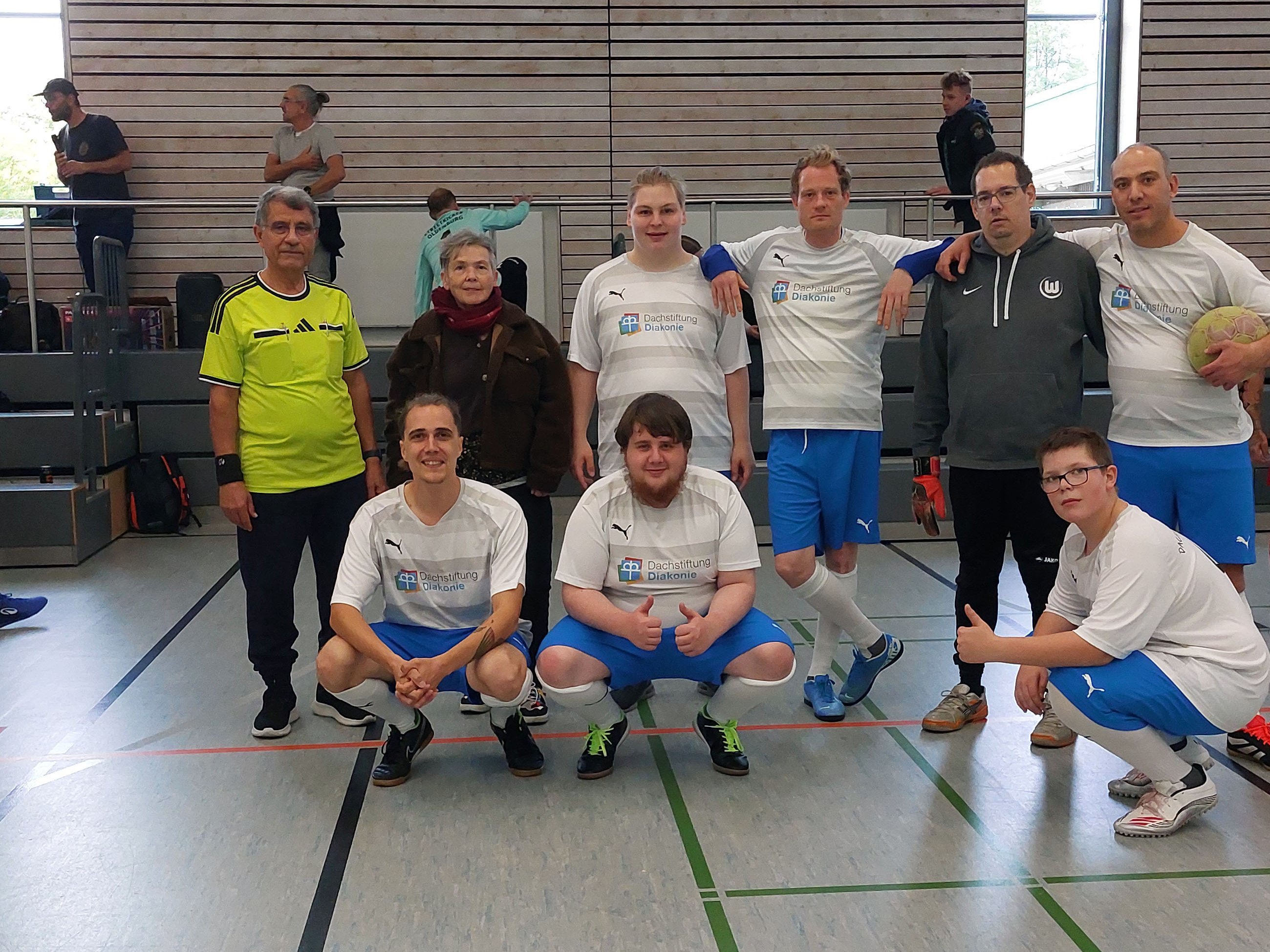 Gruppenfoto der Mannschaft der Diakonischen Betriebe Kästorf in einer Sporthalle. Neun Personen posieren in zwei Reihen, vier davon hocken vorne. Die meisten Spieler tragen weiße Fußballtrikots mit dem Logo der Dachstiftung Diakonie und blaue Hosen. Rechts hält ein Spieler einen Fußball im Arm.
