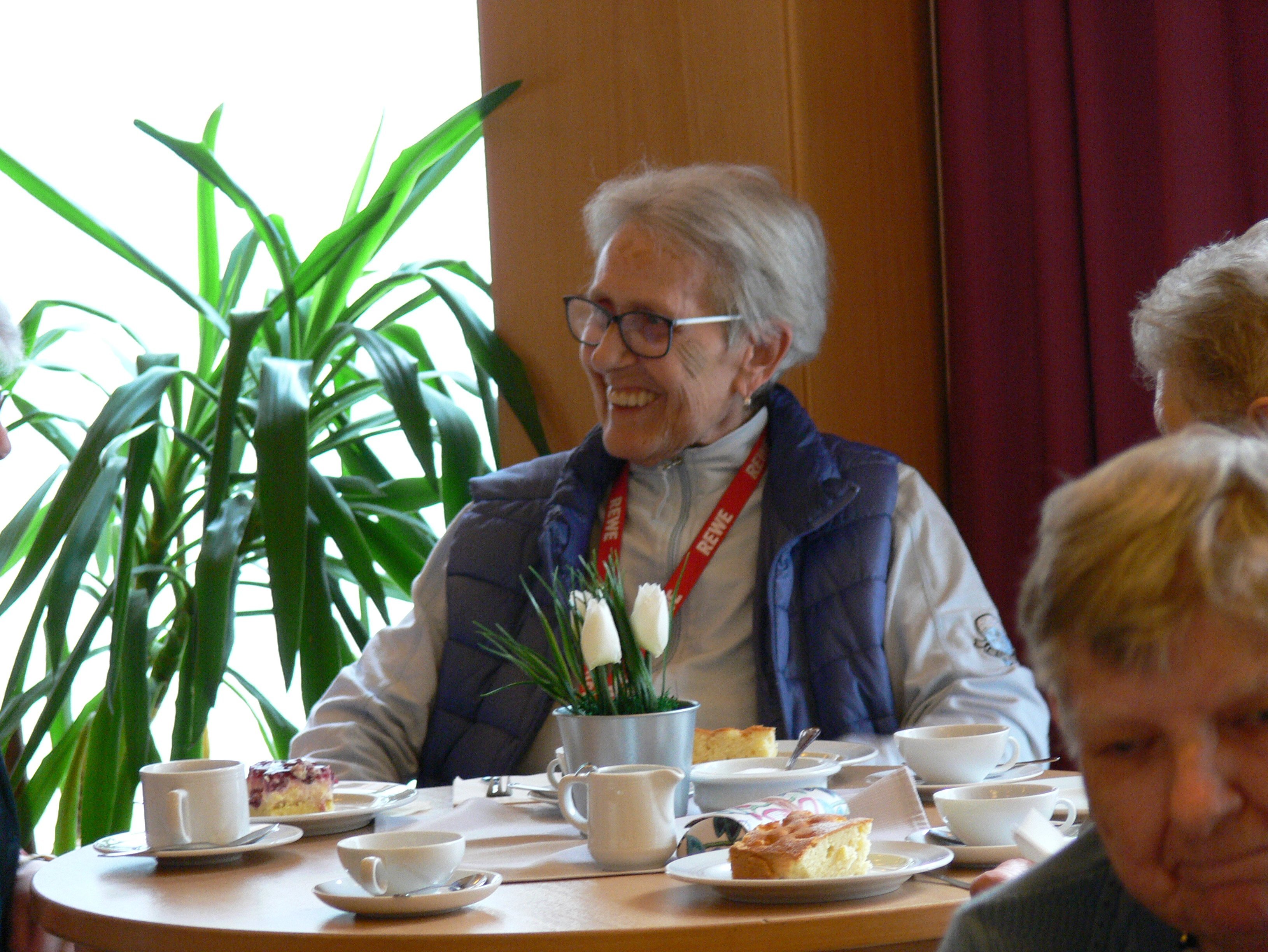 Eine ältere Frau mit weißem Haar und Brille lächelt an einem Kaffeetisch mit Kuchen, Geschirr und einem kleinen Blumengesteck.