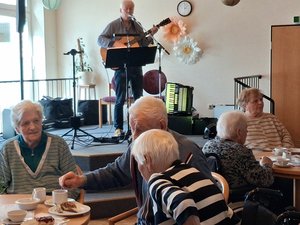 Musiker Peter Focken singt auf einer kleinen Bühne und spielt Gitarre, während Seniorinnen und Senioren an gedeckten Kaffeetischen sitzen.