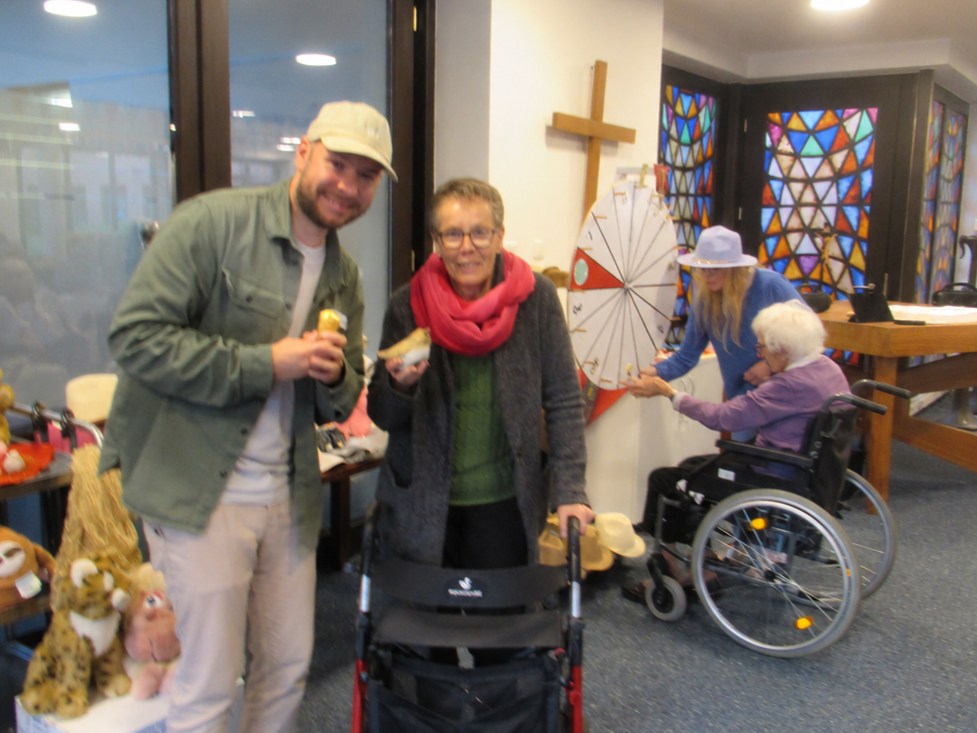Ein lächelnder Mann mit heller Kappe steht neben einer Frau mit rotem Schal und Rollator. Beide halten eine kleine Leckerei in der Hand. Im Hintergrund versucht eine ältere Frau im Rollstuhl ihr Glück am Glücksrad, begleitet von einer Frau mit blauem Oberteil und hellem Hut.