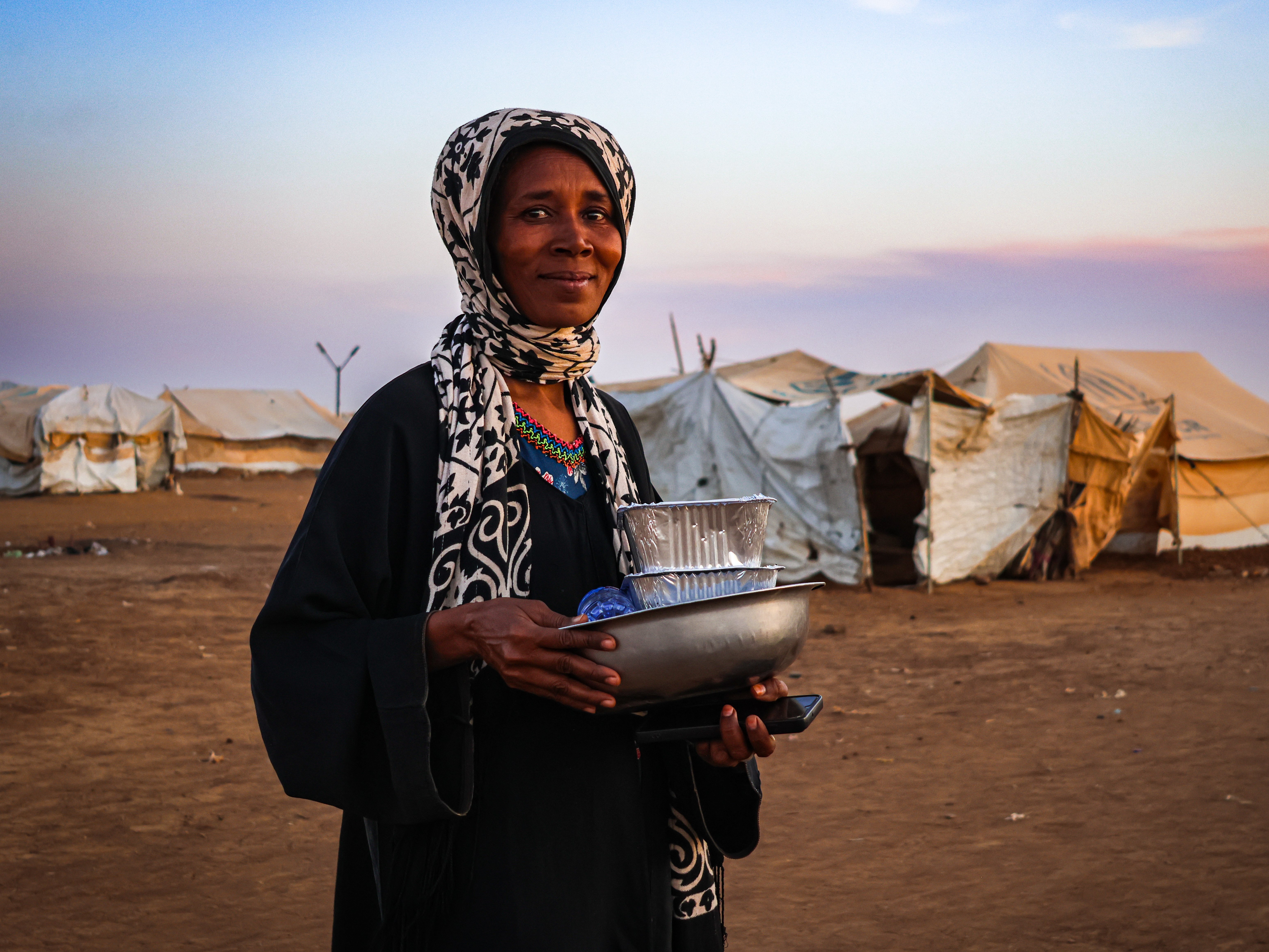 Eine Frau in einem Flüchtlingscamp im Sudan hält Schüsseln und Lebensmittelbehälter in den Händen. Hinter ihr stehen provisorische Zelte. 