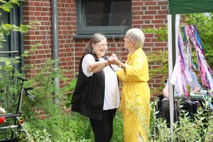 Zwei ältere Frauen tanzen lächelnd miteinander im Garten des Elisabethstifts. Eine trägt eine schwarze Weste über einer weißen Bluse, die andere ein leuchtend gelbes Kleid. Sie halten sich an den Händen. Im Hintergrund sind eine Backsteinwand, grüne Pflanzen und bunte Dekobänder zu sehen.
