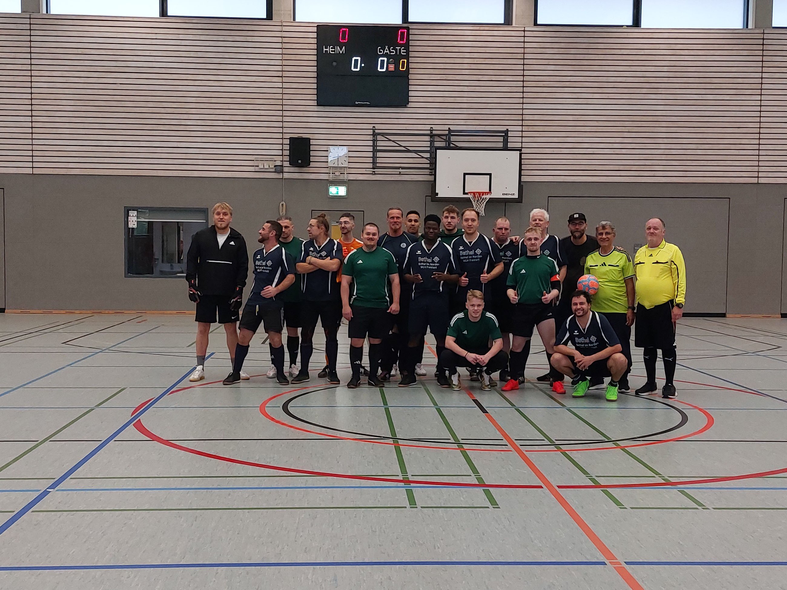 Gruppenfoto der beiden Finalmannschaften Bethel Freistatt und Karl-Jaspers-Klinik (KJK) beim 1. Moin Cup in Oldenburg. Die Spieler stehen und knien gemeinsam in der Mitte einer Sporthalle. Sie tragen überwiegend dunkelblaue und dunkelgrüne Fußballtrikots. Rechts stehen zwei Personen in gelben Schiedsrichtertrikots.