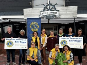 Gruppenfoto vor den Weserterrassen: Mehrere Frauen und ein Mann stehen und knien vor dem Eingang. Einige Frauen tragen gelbe Lions-Westen und halten Kleidungsstücke in den Händen. Zwei Personen halten Spendenschecks des Lions Clubs „Emy Rogge“ in die Kamera.
