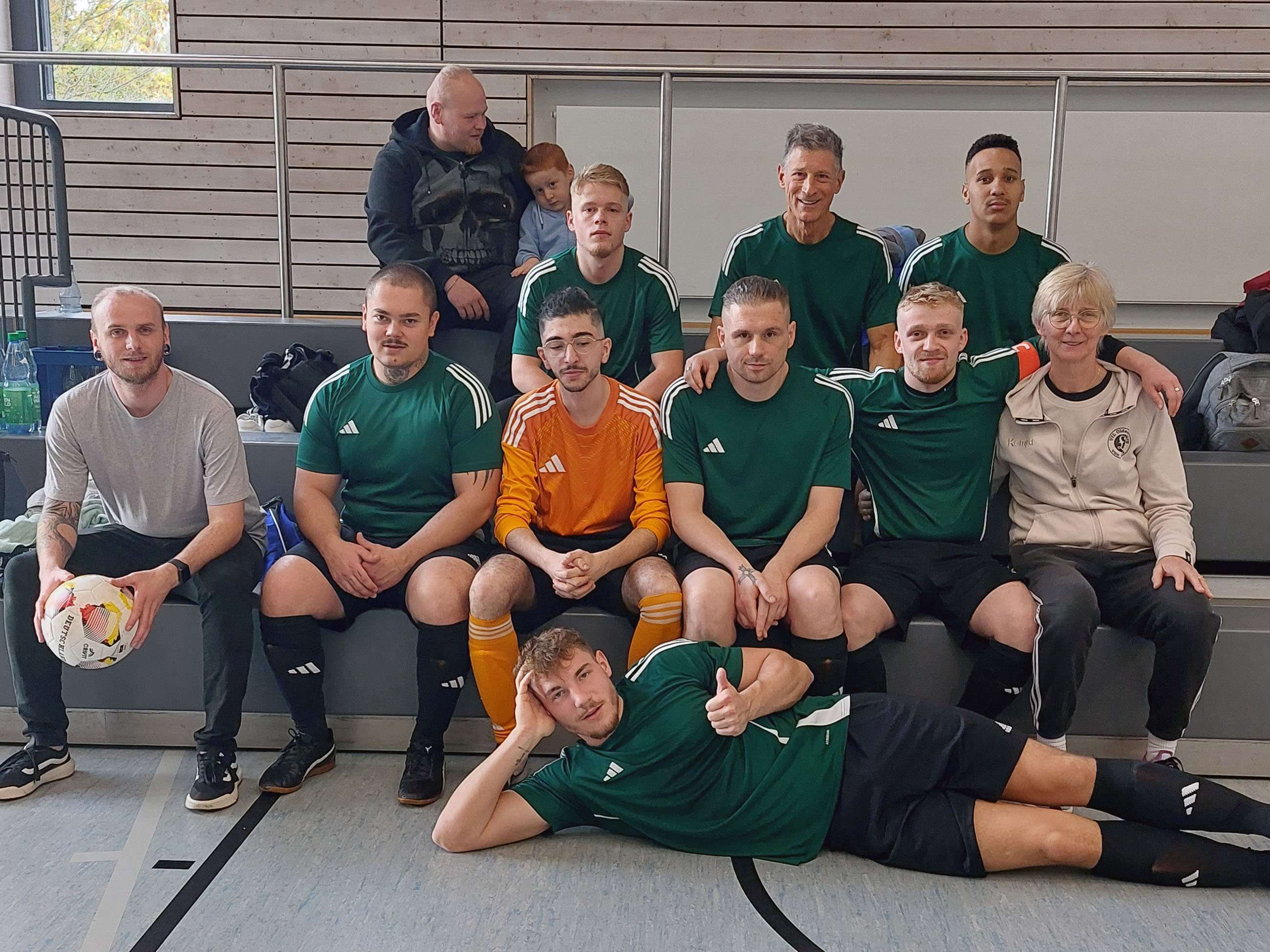 Gruppenfoto der Mannschaft der Karl-Jaspers-Klinik in einer Sporthalle. Die meisten Spieler tragen dunkelgrüne Fußballtrikots, der Torwart ein orangefarbenes Trikot. Mehrere Personen sitzen auf einer Bank, ein Spieler liegt davor auf der Seite und zeigt den Daumen nach oben.