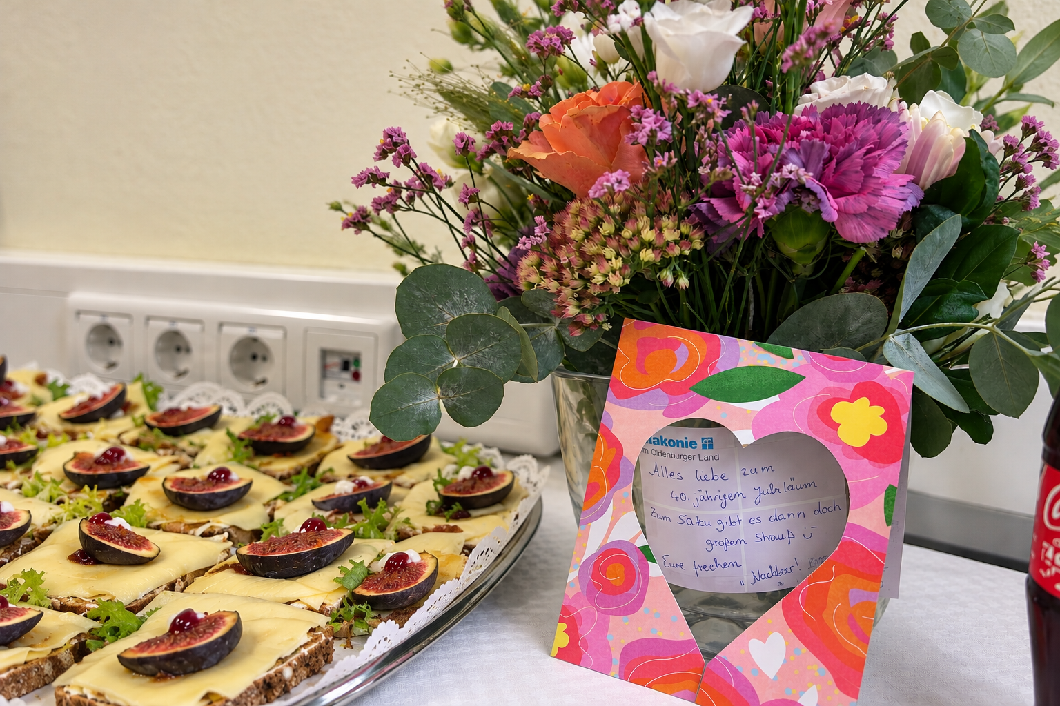 Bunter Blumenstrauß mit rosa, weißen und orangefarbenen Blüten neben einer Glückwunschkarte zum 40-jährigen Jubiläum. Im Vordergrund stehen belegte Brote mit Käse und Feigen.