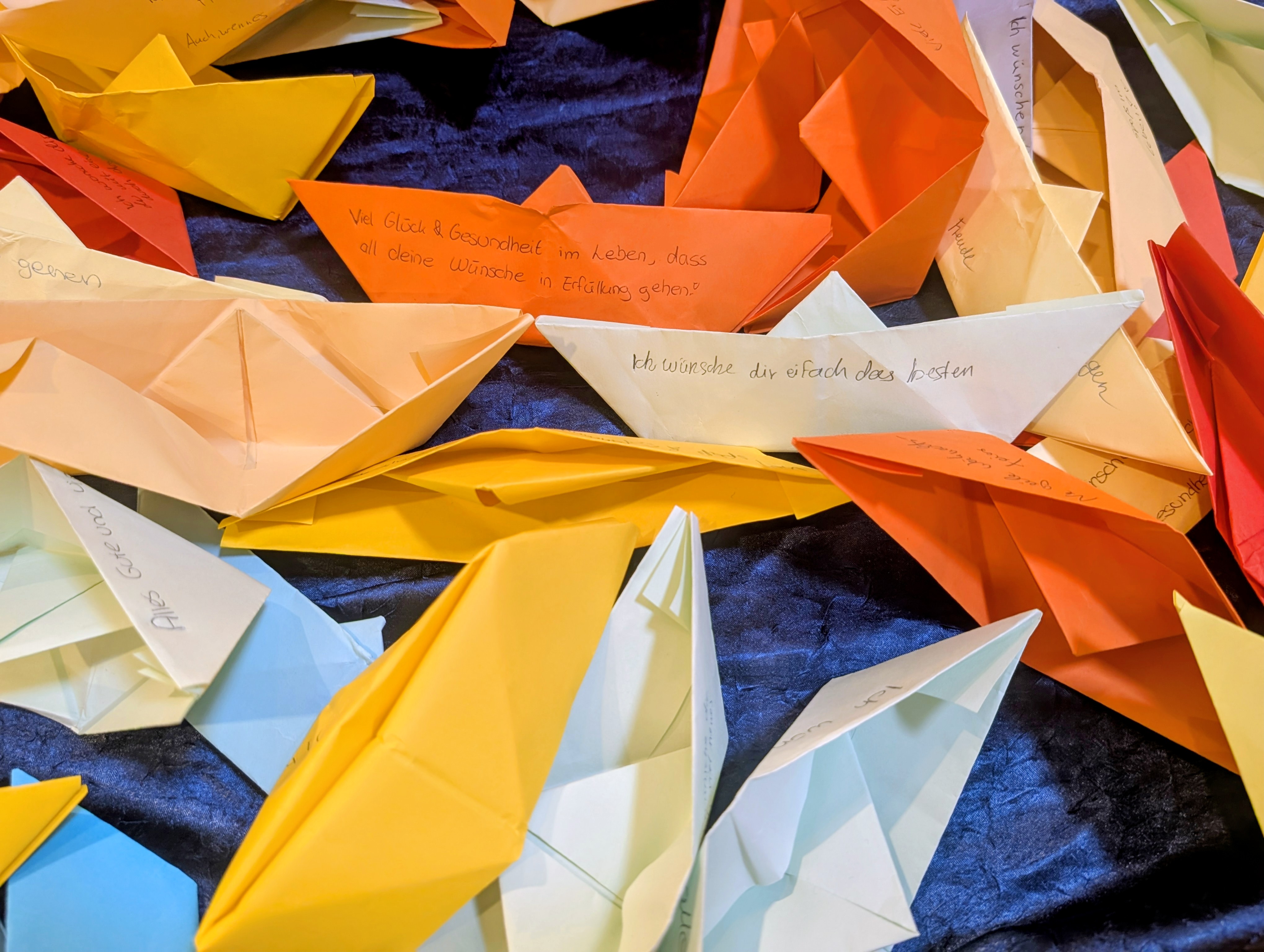 Viele bunte, mit handgeschriebenen Wünschen versehene Papierschiffchen liegen auf einem dunkelblauen Stoff.