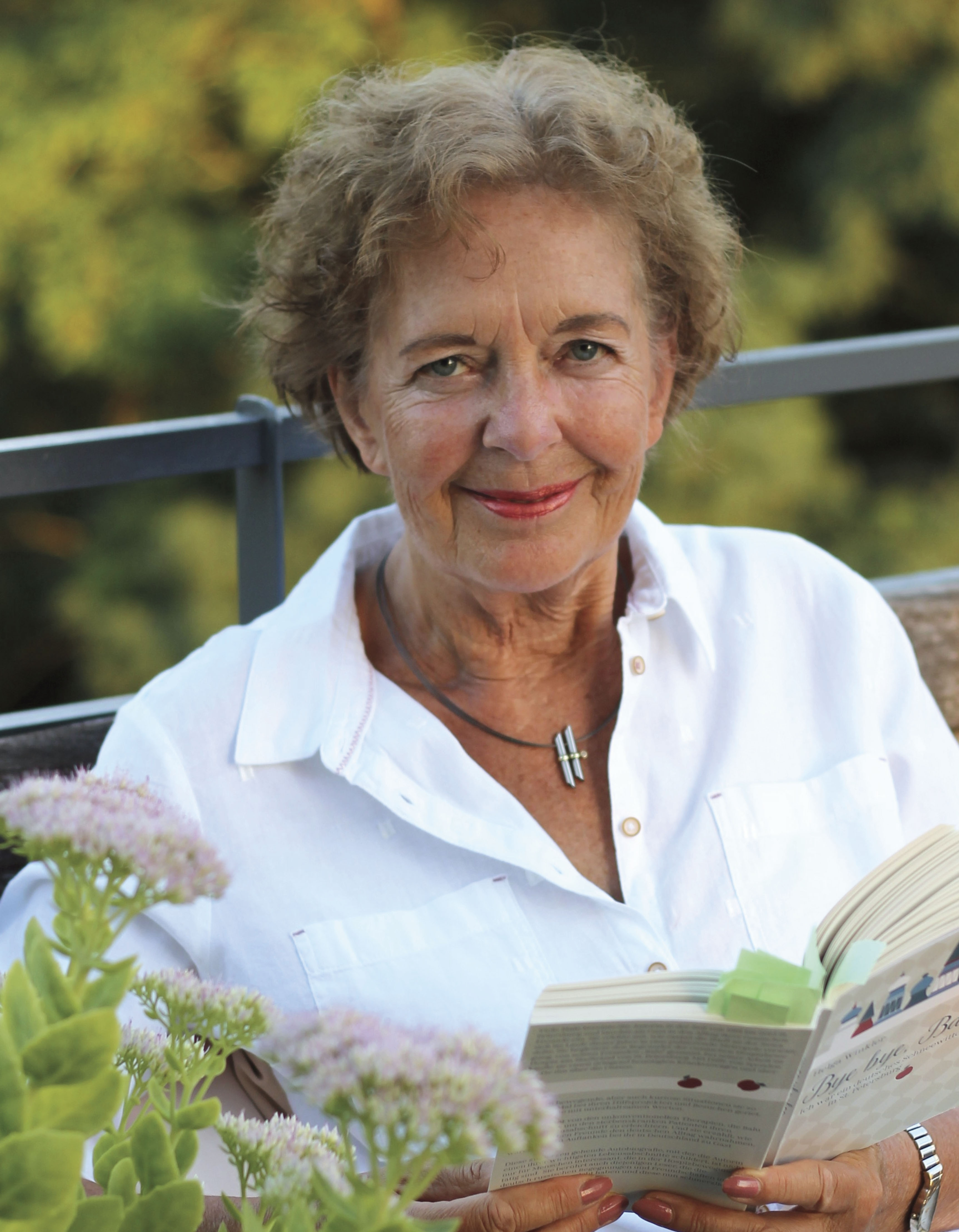 Helga Winkler sitzt im Freien, lächelt in die Kamera und hält ein aufgeschlagenes Buch in den Händen. Sie trägt eine weiße Bluse, im Vordergrund sind Blumen, im Hintergrund grünes Blattwerk.