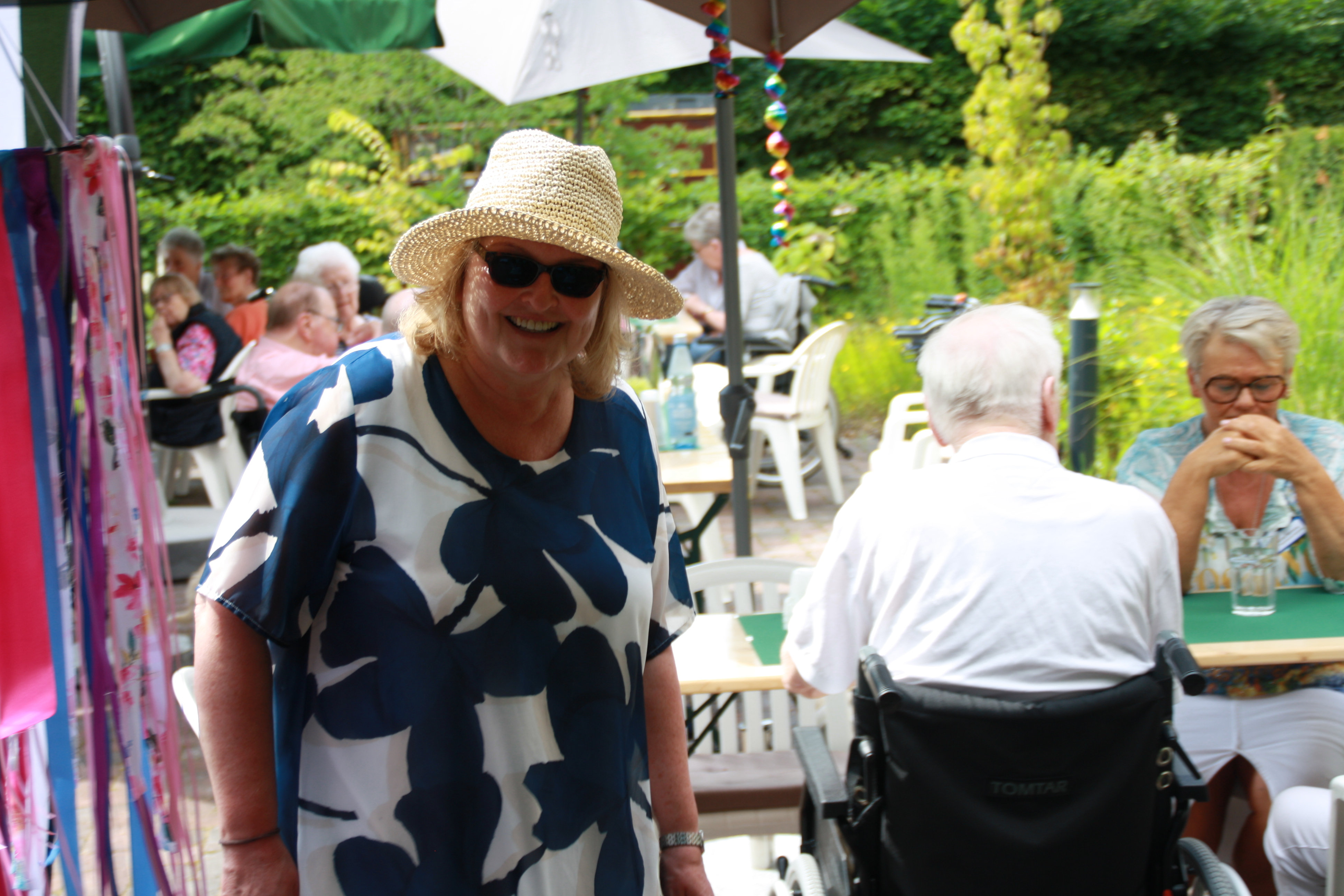 Oberin Frau Schumann steht lächelnd im Garten des Elisabethstifts. Sie trägt einen Strohhut, eine Sonnenbrille und ein blau-weiß gemustertes Kleid. Im Hintergrund sitzen Gäste des Sommerfestes an Tischen unter Sonnenschirmen.
